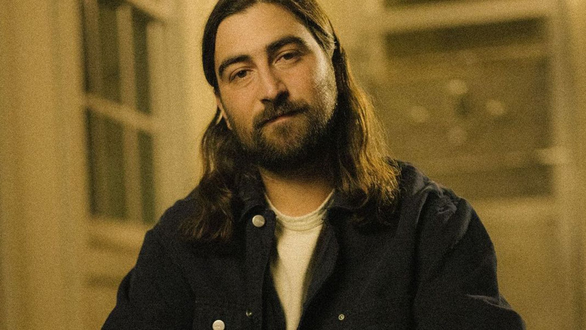 Porträtfoto von Noah Kahan mit langen Haaren und Bart, sitzend in einem Innenraum mit warmem, gedämpftem Licht und unscharfem Hintergrund mit Fensterm