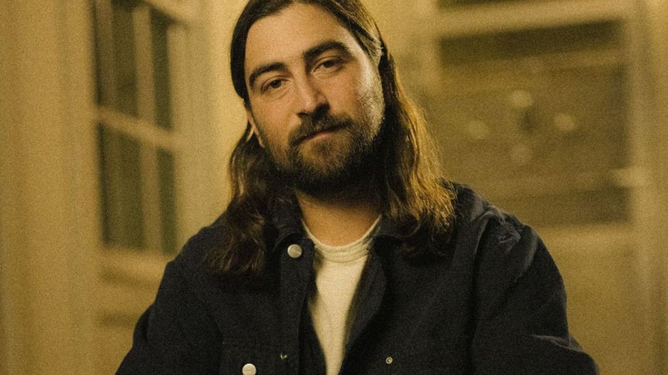 Porträtfoto von Noah Kahan mit langen Haaren und Bart, sitzend in einem Innenraum mit warmem, gedämpftem Licht und unscharfem Hintergrund mit Fensterm