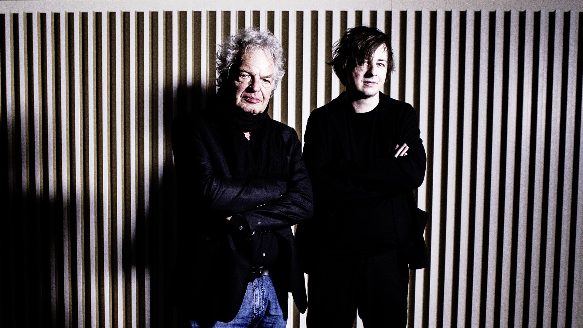 Michael Wollny und Joachim Kühn vor einer Wand mit vertikalen Lamellen, beide in dunkler Kleidung mit verschränkten Armen vor kontrastreicher Beleuchtung
