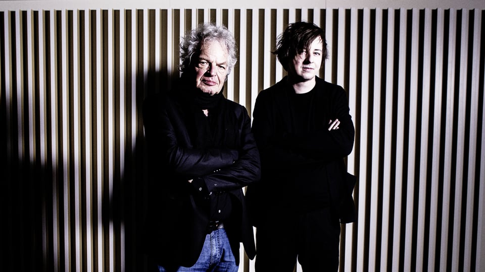 Michael Wollny und Joachim Kühn vor einer Wand mit vertikalen Lamellen, beide in dunkler Kleidung mit verschränkten Armen vor kontrastreicher Beleuchtung