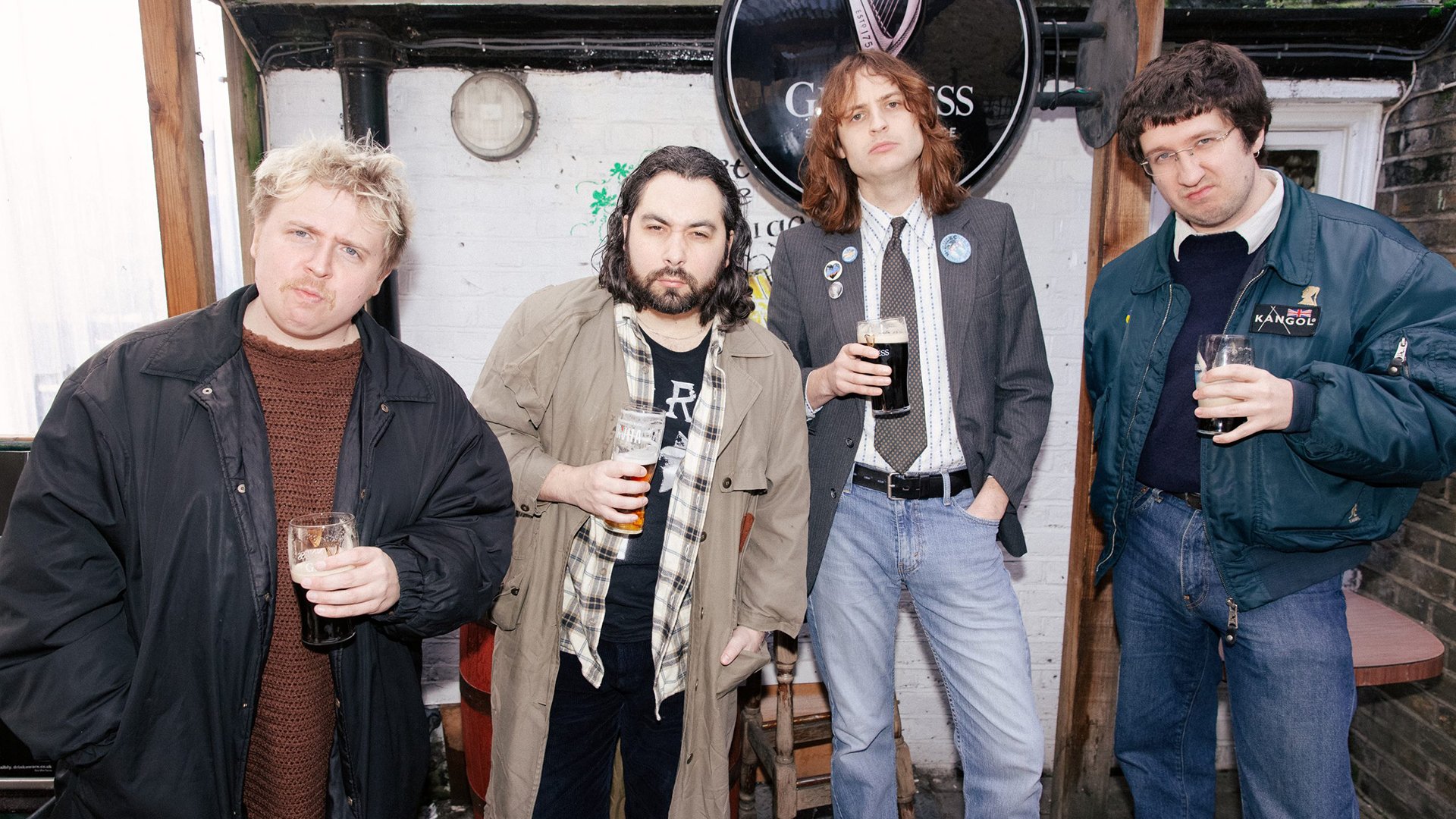 vier Personen stehen in legerer Kleidung vor einer weißen Außenwand mit rundem Guinness‑Schild, jede Person hält ein Getränk in der Hand. Tageslicht, informelle Atmosphäre; Motiv als Promo‑ und Eventbild der Band THE TUBS.