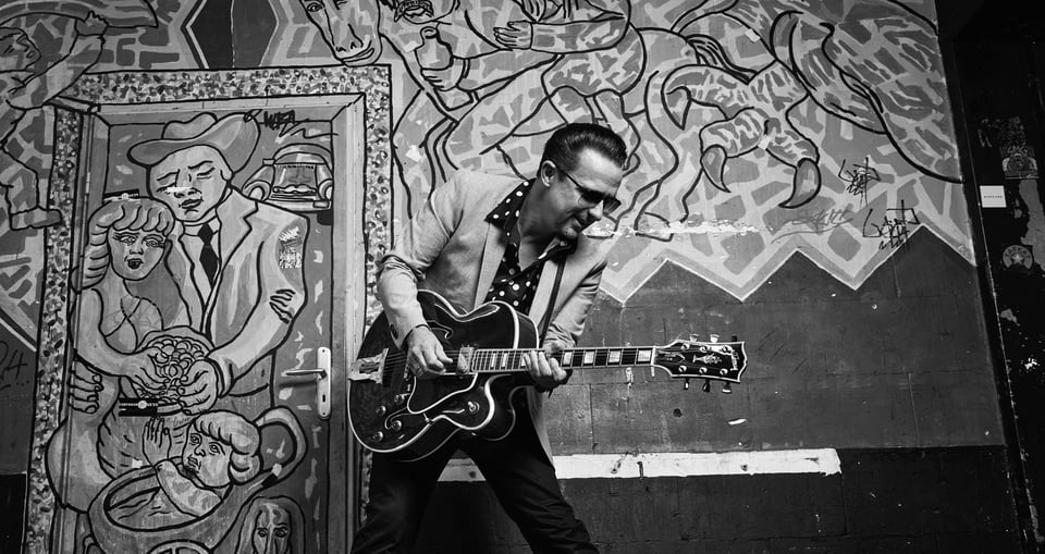 schwarz weiss Bild von Mann mit Gitarre vor Wand und Türe mit Zeichnungen