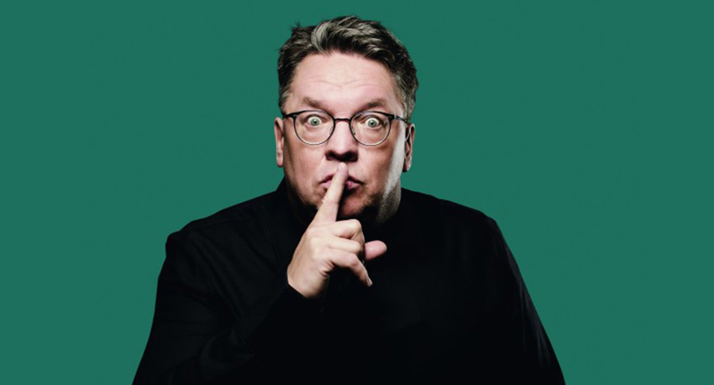 Der Künstler in schwarzem Pullover, den Zeigefinger an den Mund haltend, vor grünem Hintergrund