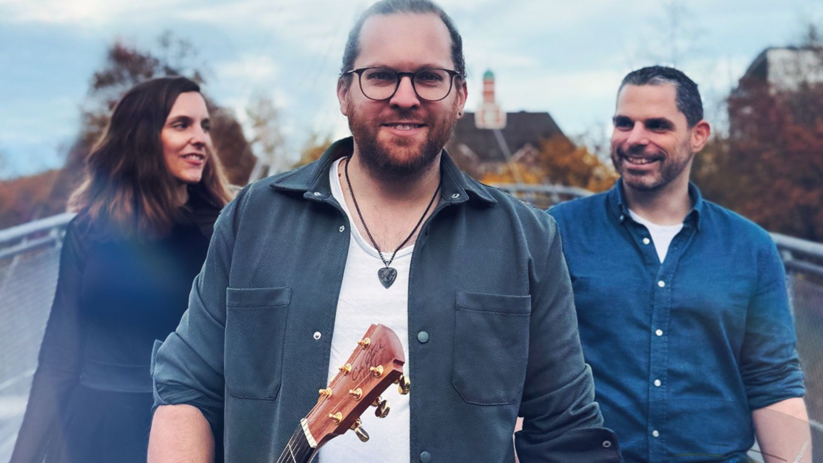 Drei Personen gehen über eine Fußgängerbrücke im Freien; die Person in der Mitte hält eine Gitarre, die beiden anderen gehen seitlich versetzt, herbstliche Bäume im Hintergrund