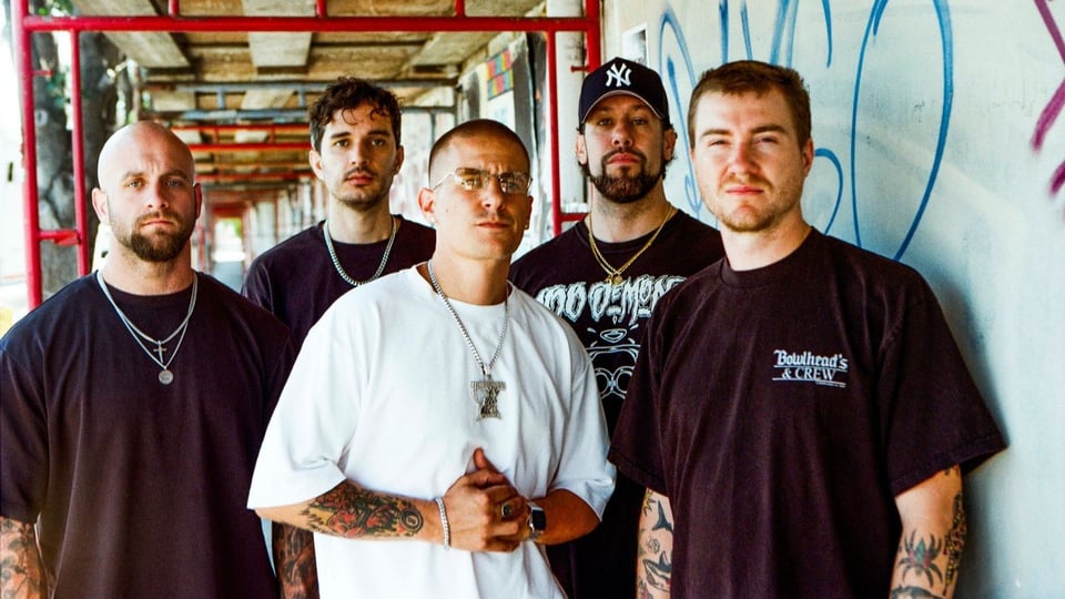 Bandfoto von Pain Of Truth in dunkler Freizeitkleidung vor einer betonierten Passage mit roten Gerüststangen und Graffiti, eine Person im weißen T-Shirt im Vordergrund sowie eine Person mit Baseballkappe im Hintergrund.
