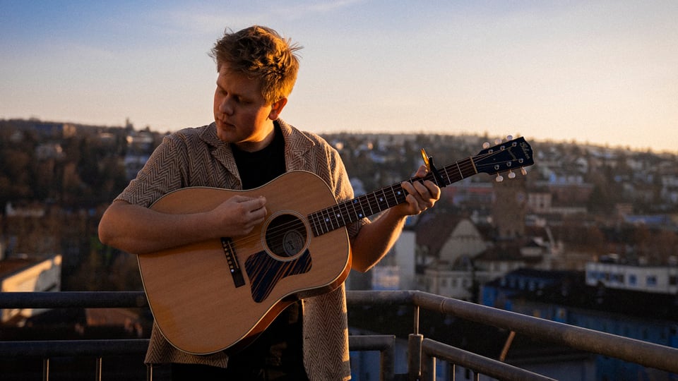 Eine Person spielt eine akustische Gitarre auf einem Balkon oder einer Dachterrasse mit Blick über eine Stadt bei warmem Abendlicht.