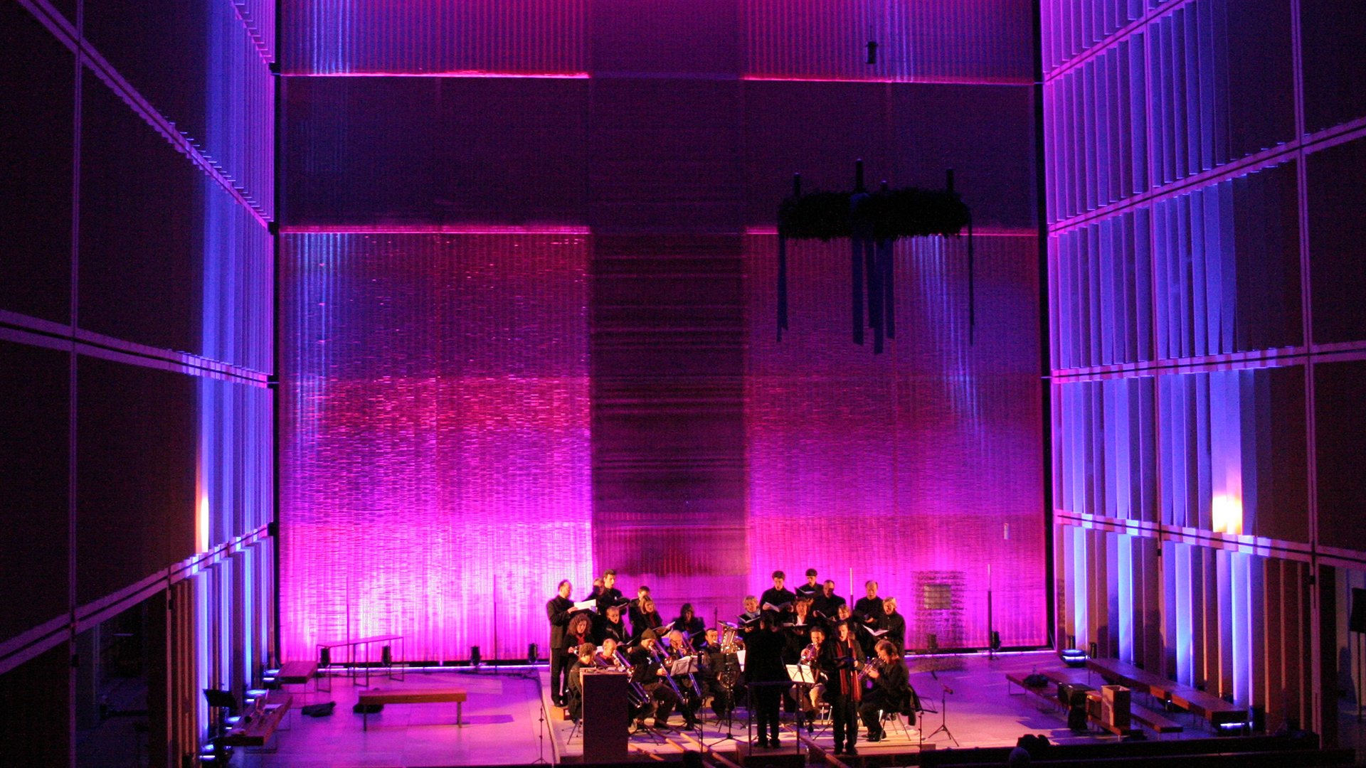 Bühnenfoto von Ascending Voices mit Chor und Kammerensemble auf einer Bühne der Herz-Jesu-Kirche, vor einer großflächig in Pink und Violett beleuchteten Architekturkulisse mit hohen Wandlamellen und transparentem Vorhang im Hintergrund