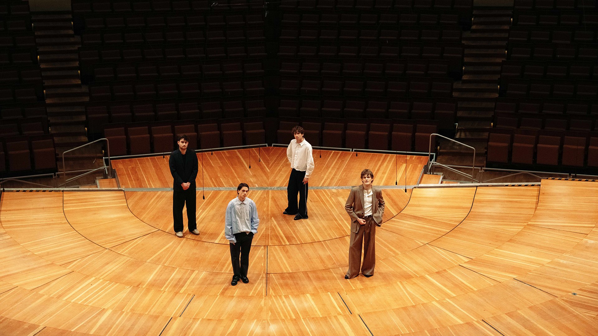 Pressefoto zu JEREMIAS & Ensemble 2026: fünf Personen stehen auf einer großen runden Holz‑Bühne in einem leeren Konzertsaal mit ansteigenden Sitzreihen im Hintergrund. Weite Perspektive, warme Holzoberflächen und klare Linien; Motiv zur Ensemble‑Tour der Indie‑Pop‑Band JEREMIAS.