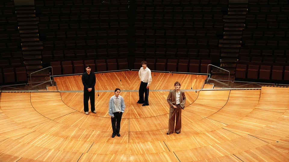 Pressefoto zu JEREMIAS & Ensemble 2026: fünf Personen stehen auf einer großen runden Holz‑Bühne in einem leeren Konzertsaal mit ansteigenden Sitzreihen im Hintergrund. Weite Perspektive, warme Holzoberflächen und klare Linien; Motiv zur Ensemble‑Tour der Indie‑Pop‑Band JEREMIAS.