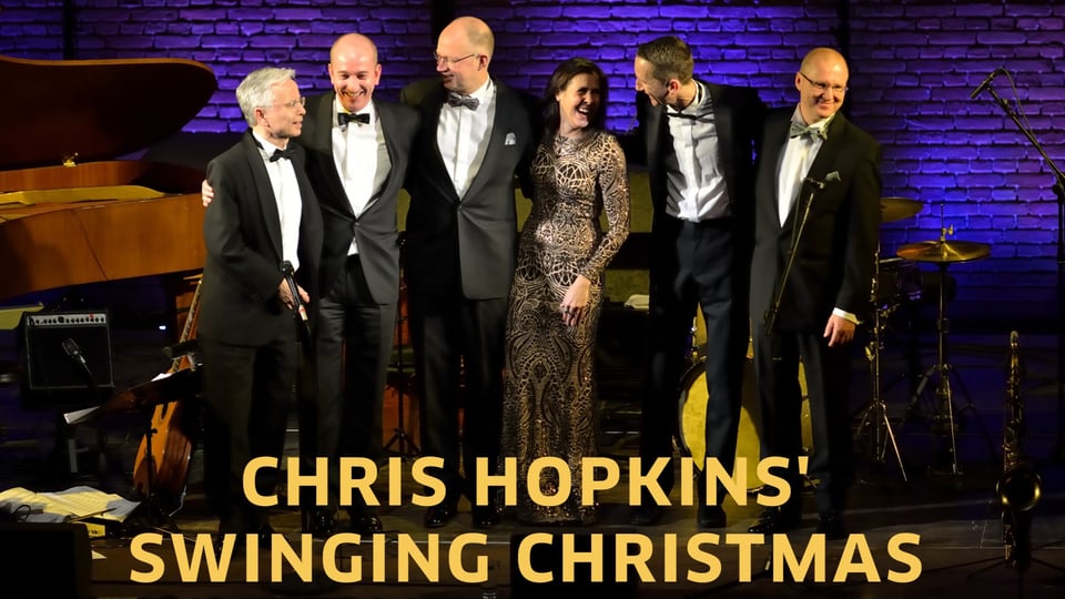 Sechs elegant gekleidete Musikerinnen und Musiker stehen Arm in Arm auf einer Bühne vor Instrumenten und Backsteinwand, darunter der Schriftzug Chris Hopkins – Swinging Christmas  in gelb