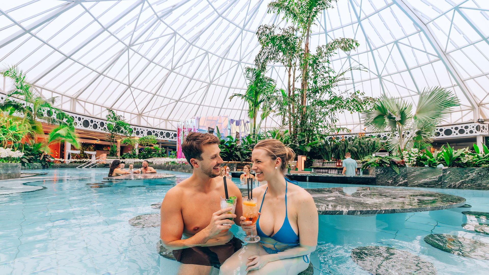 Zwei Personen sitzen am Rand eines Innenpools unter einer großen Glaskuppel. Vor ihnen stehen zwei bunte Cocktails