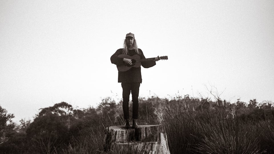 ein schwarz-weiß Foto des Künstlers mit Gitarre auf einem Baumstumpf stehend 