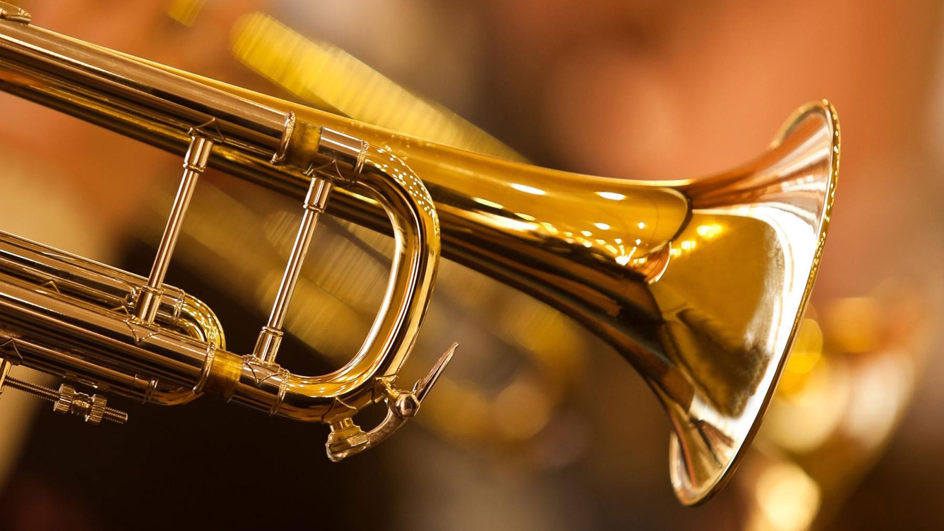 Nahaufnahme einer goldenen Trompete mit Ventilen und Schalltrichter vor warmem, unscharfem Hintergrund mit Lichteffekten in goldenen und orangefarbenen Tönen.