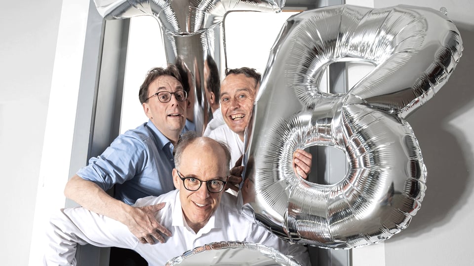 Drei Personen stehen in einem Türrahmen zwischen silbernen Folienballons in Form der Buchstaben T B und C und blicken in die Kamera