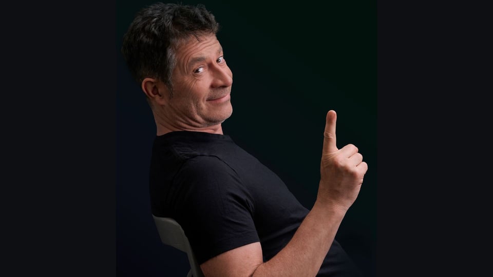 Rolf Miller auf einem Stuhl in schwarzem T-Shirt und erhobenem Zeigefinger blickt seitlich in die Kamera