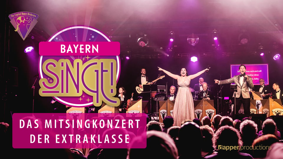 Bühnenfoto eines Mitsingkonzerts mit Big Band, Solisten und Publikum vor violetter Beleuchtung. Im Vordergrund steht der Schriftzug Bayern Singt!