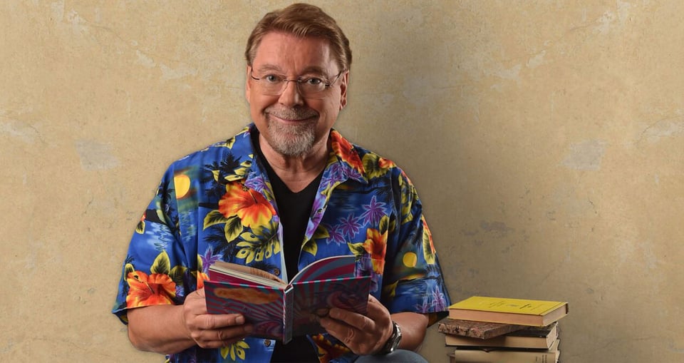 Foto des Künstlers Jürgen von der Lippe mit einem Hawaiihemd und einem Buch in den Händen vor einer gelben Wand