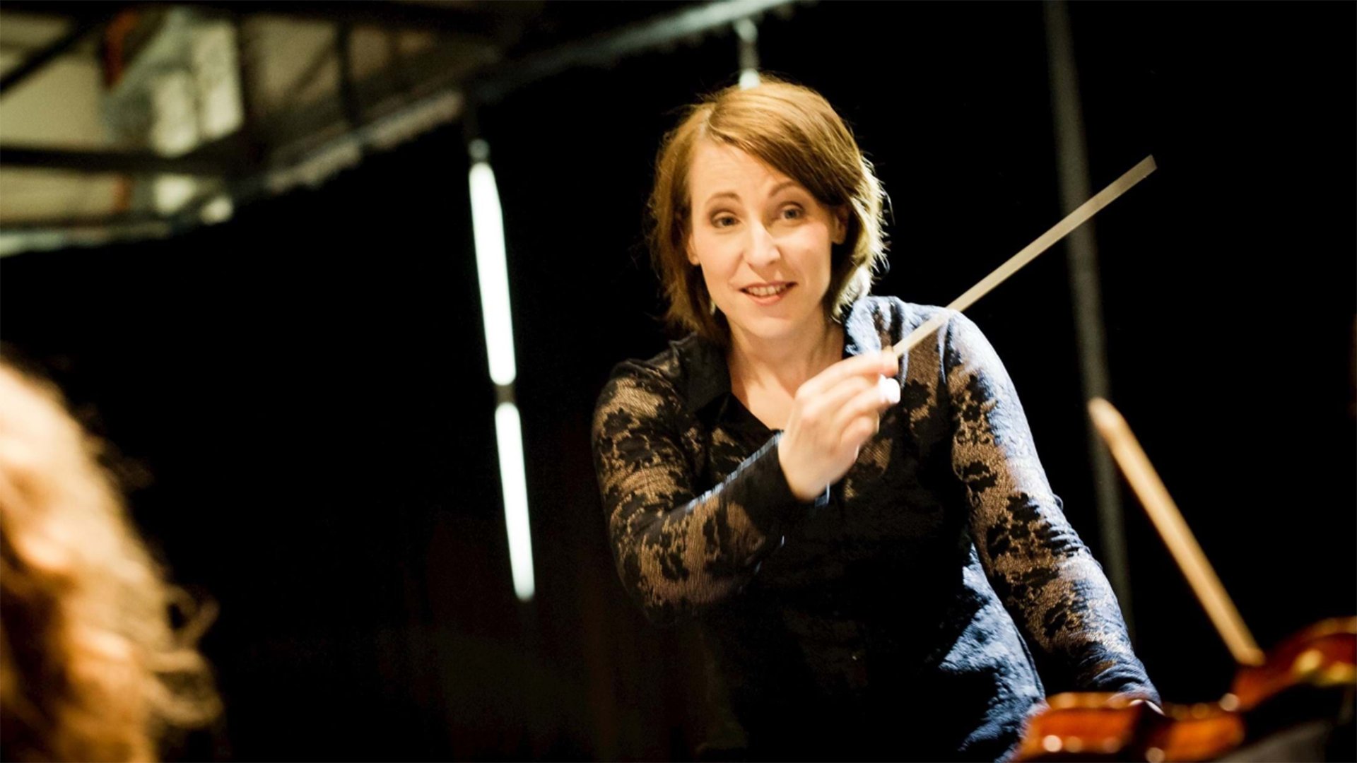 Bühnenfoto von Carolin Nordmeyer mit erhobenem Dirigierstab vor einem Orchester, in schwarzer Konzertkleidung und warmem Bühnenlicht, zwischen Streichinstrumenten auf einer Konzertbühne