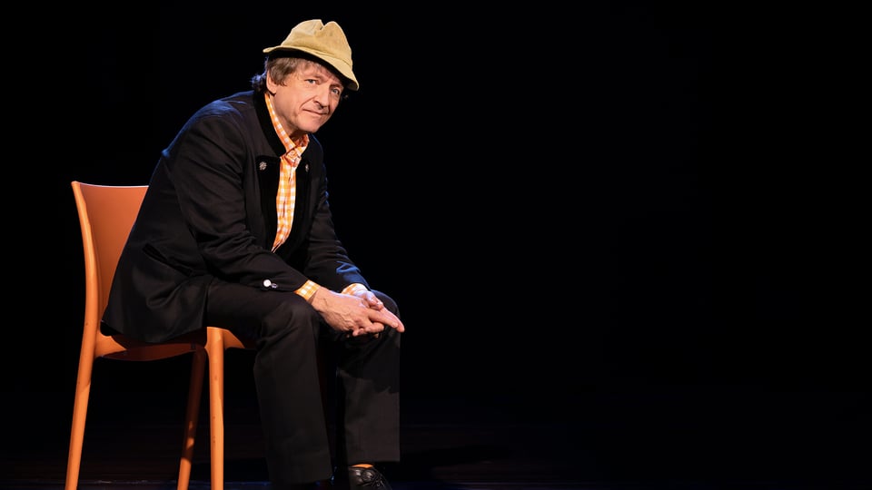 Erwin Pelzig sitzt auf einer Bühne auf einem Stuhl und guckt schräg in die Kamera