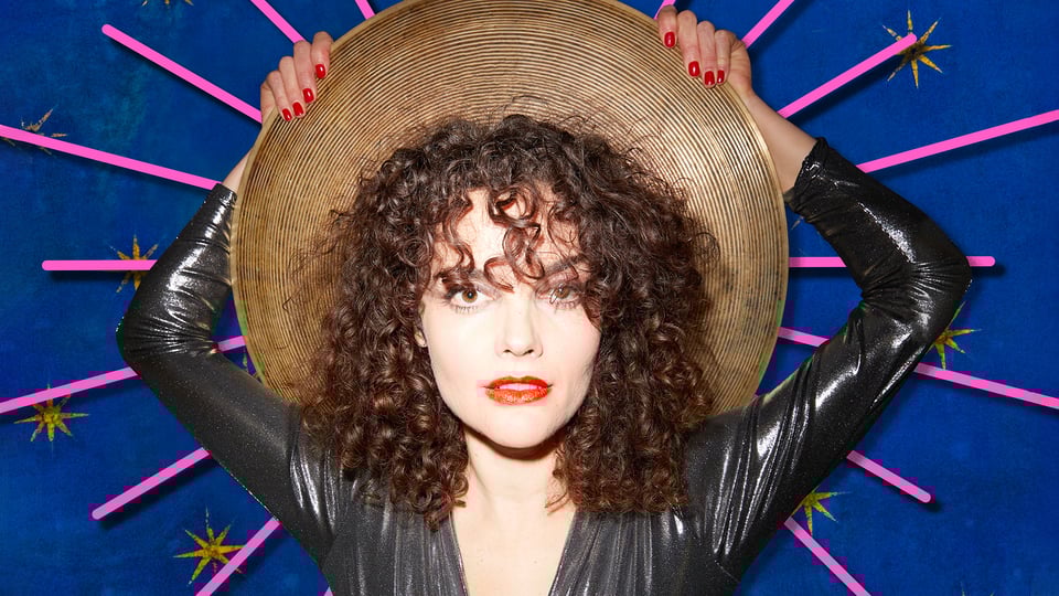 Ein Foto von Maria de Val mit einer Art Hut in den Händen vor blauem-pinker Grafik