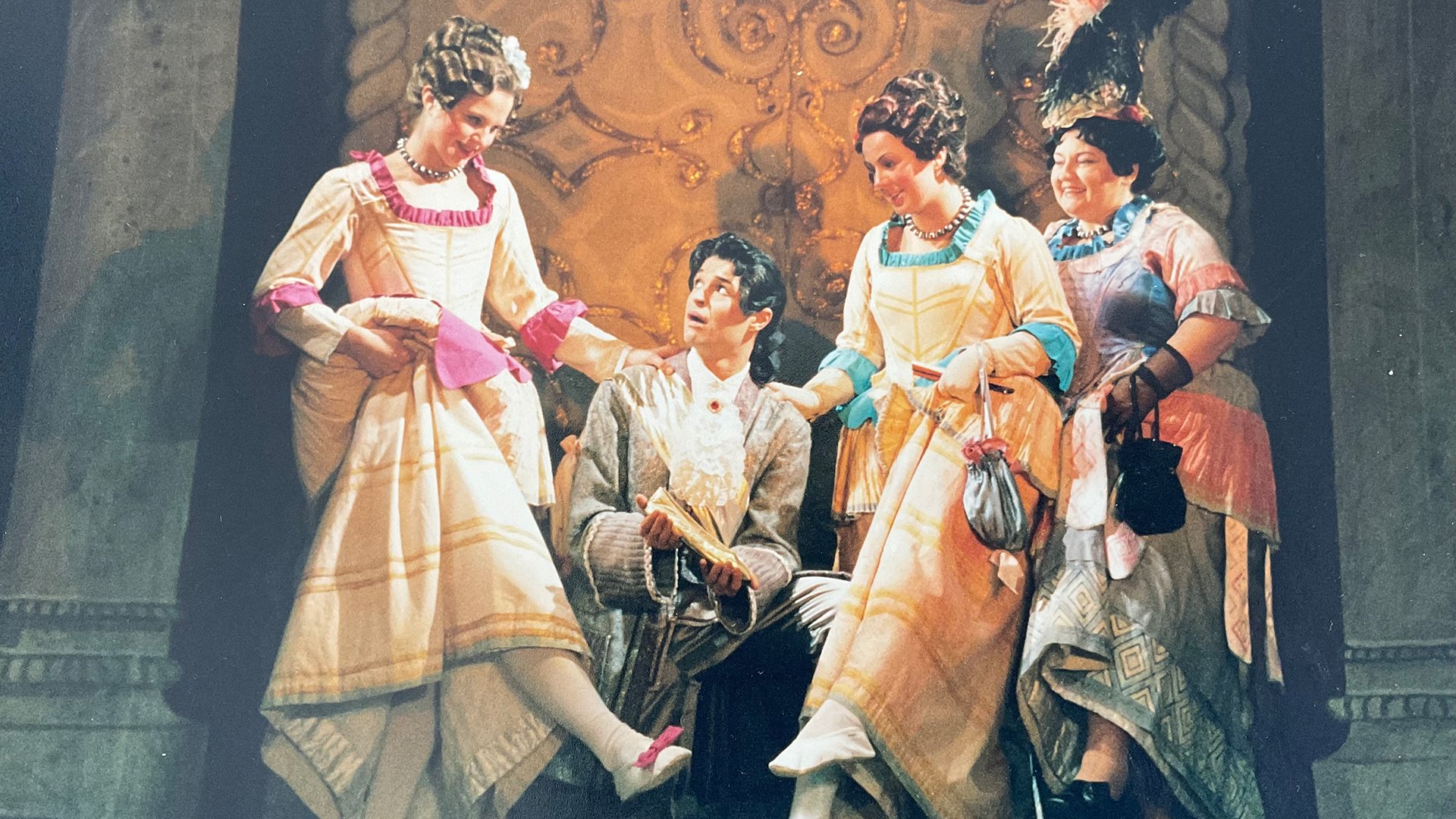 Historische Theaterszene mit vier Darstellenden in barocken Kostümen; drei Figuren in farbenfrohen Kleidern stehen um eine sitzende Person im silbernen Anzug, die ein Dokument hält, vor aufwendig verzierter Bühnenkulisse.