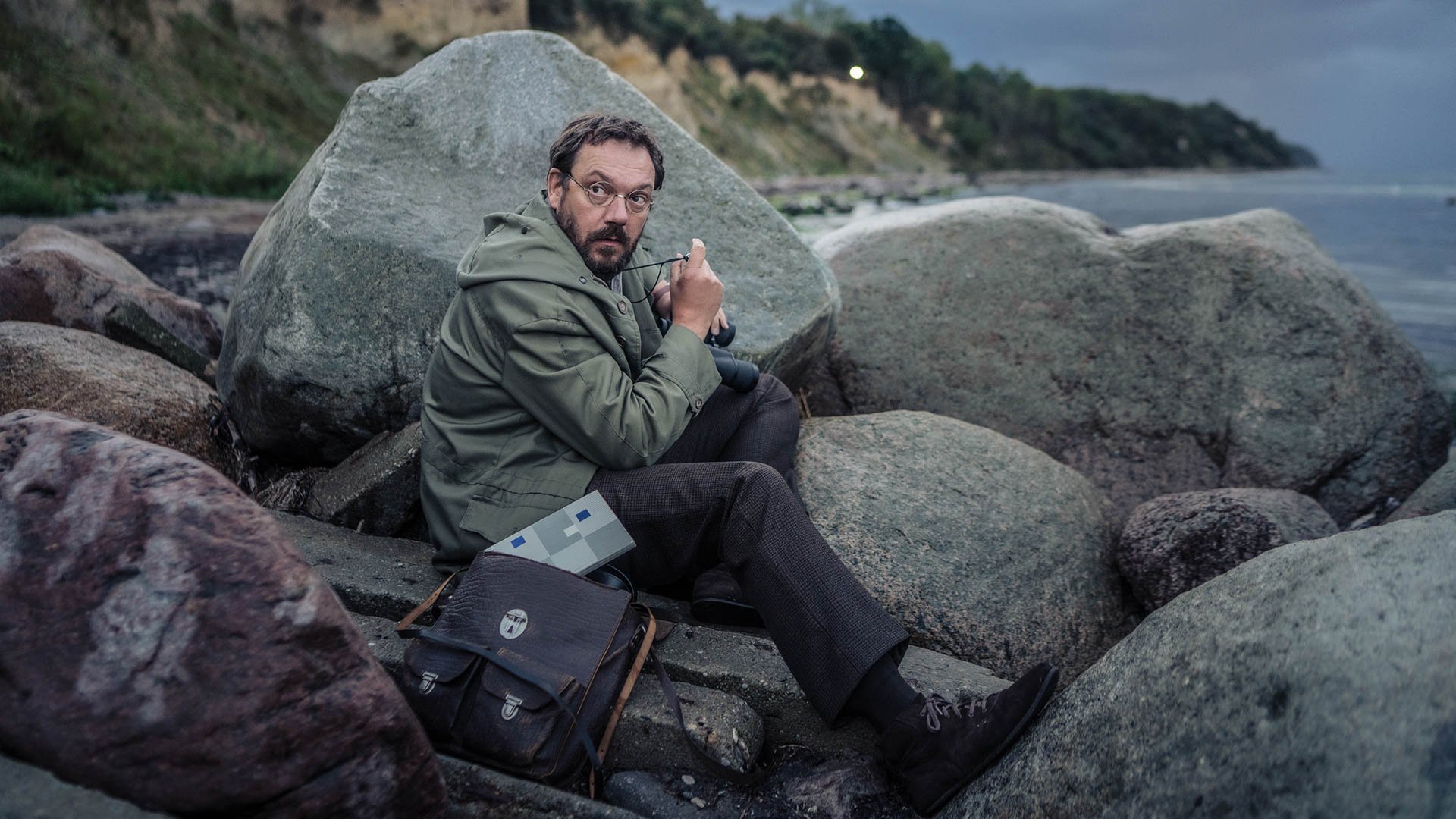 Ein Mann sitzt am Meer auf Felsen und guckt dabei nach hinten.