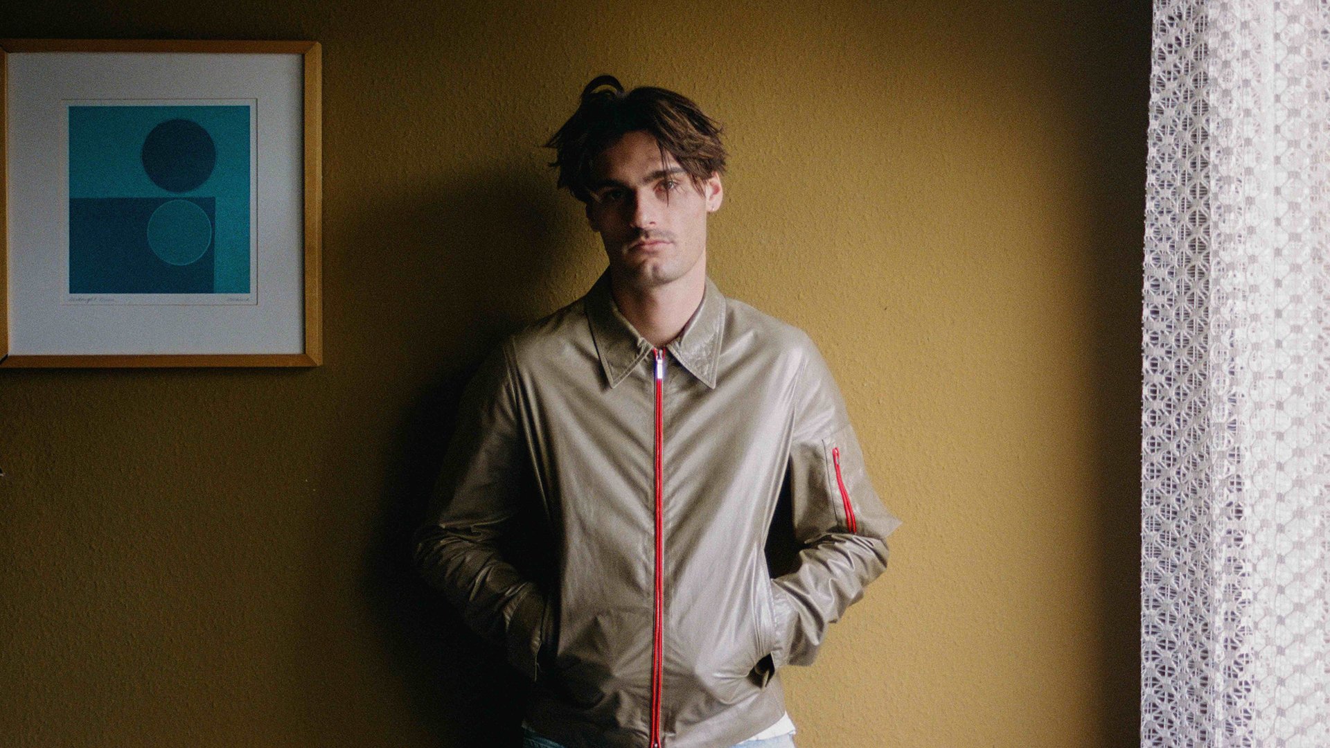 Person in beigefarbener Jacke mit rotem Reißverschluss, die mit den Händen in den Taschen vor einer ockerfarbenen Wand steht, links ein gerahmtes Bild mit geometrischen Formen und rechts ein heller Vorhang