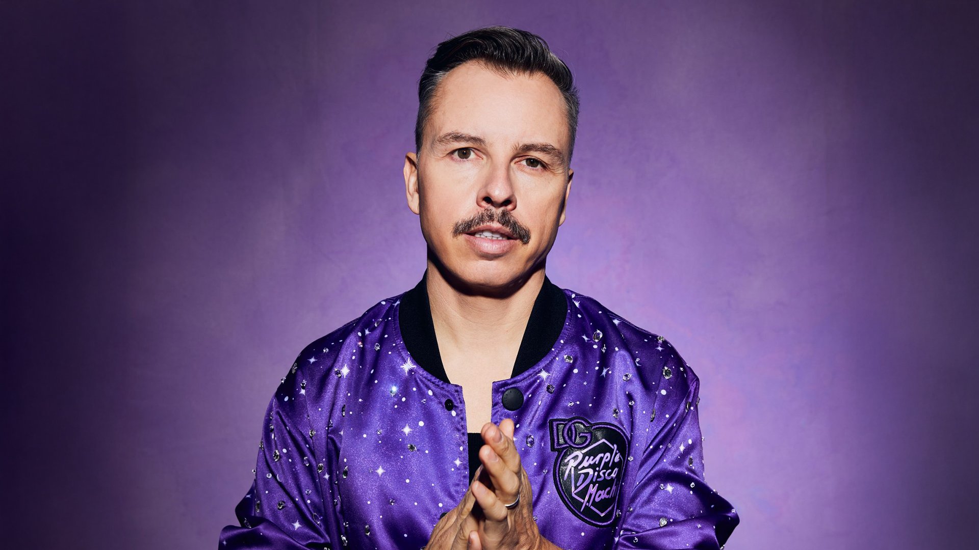 Eine Person in einer violetten Jacke mit Glitzerapplikationen blickt direkt in die Kamera, vor violettem Hintergrund