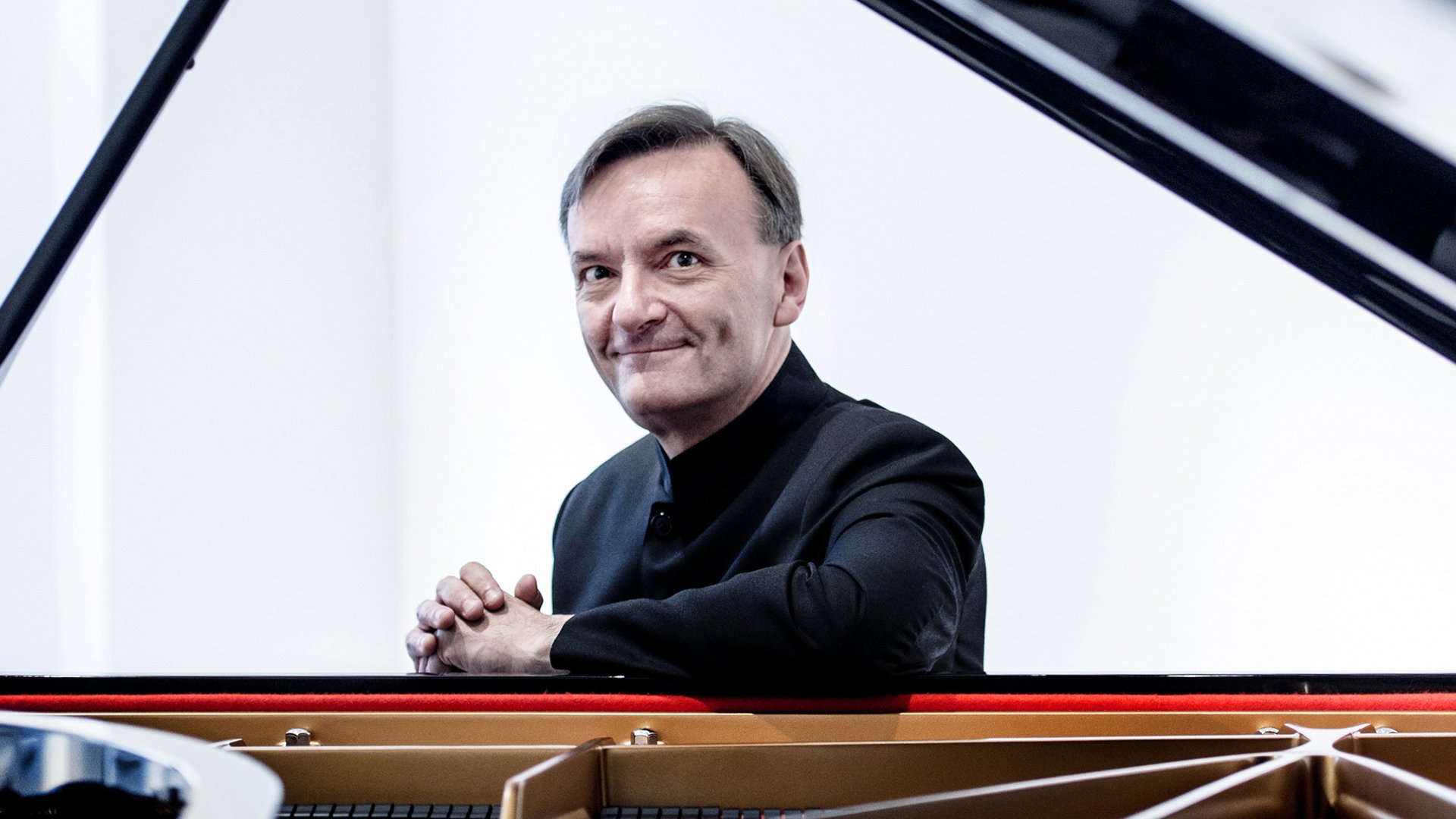 Stephen Hough im Anzug am Konzerflügel blickt lächelnd in die Kamera