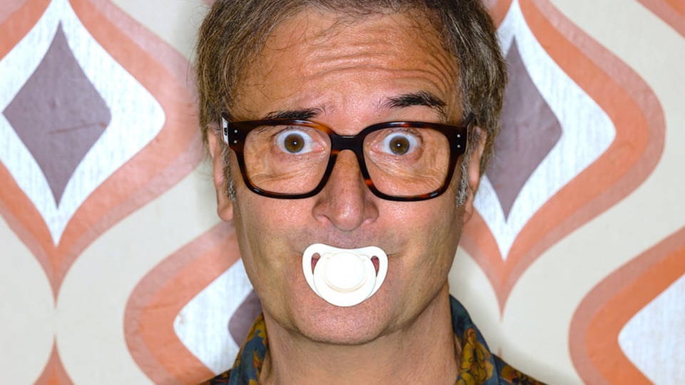 Ein Portrait von Chris Boettcher mit enem weißen Schnuller im Mund vor einer Tapete mit Muster in braun, beige und orange 