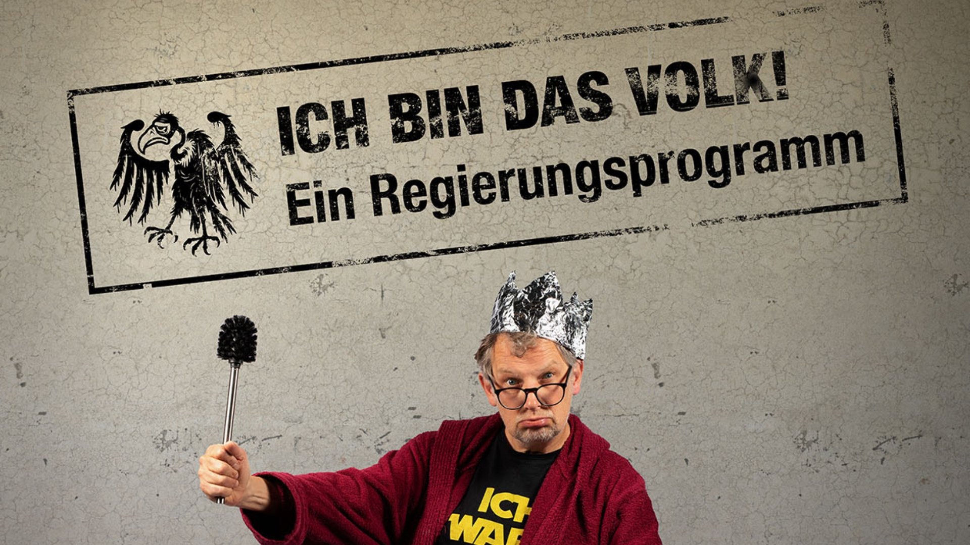 Plakatmotiv mit den Aufschriften Ich bin das Volk! und Ein Regierungsprogramm. Eine Person mit Krone aus Alufolie, Brille und rotem Bademantel hält eine Toilettenbürste wie ein Zepter vor grauem Hintergrund.