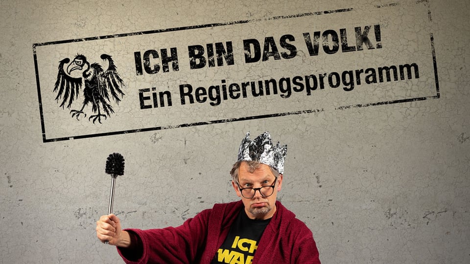 Plakatmotiv mit den Aufschriften Ich bin das Volk! und Ein Regierungsprogramm. Eine Person mit Krone aus Alufolie, Brille und rotem Bademantel hält eine Toilettenbürste wie ein Zepter vor grauem Hintergrund.
