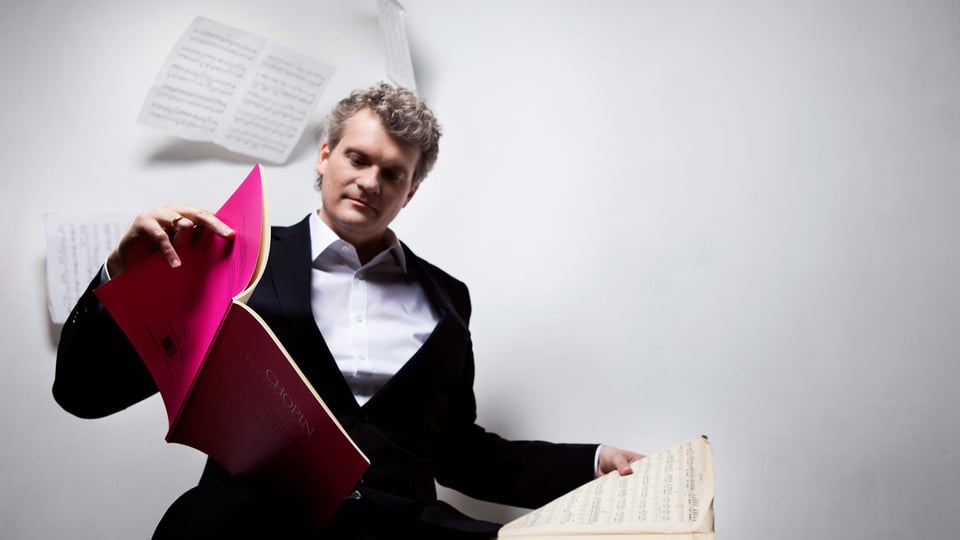 Eine Person im schwarzen Anzug blättert in Notenheften, eines mit pinkem Einband, vor hellem Hintergrund lose Notenblätter schweben im Hintergrund.