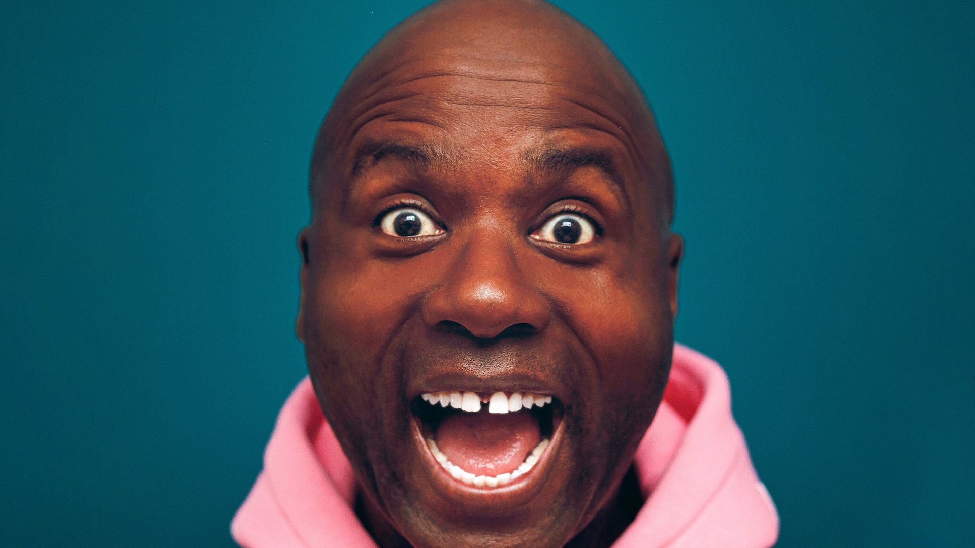 Ein Foto des lachenden Künstlers mit rosa Kapuzenpulli vor petrolfarbenem Hintergrund