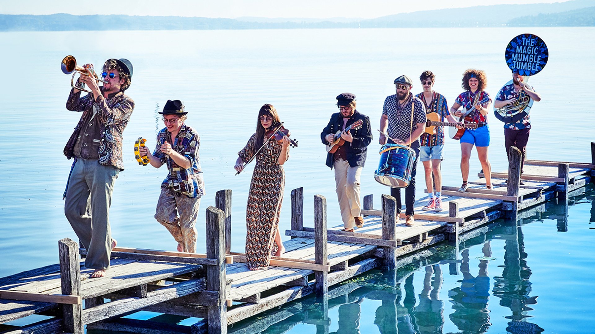 Sieben Musiker mit Instrumenten gehen auf einem Steg hintereinander