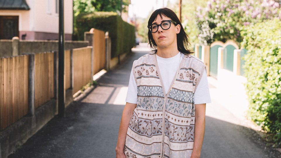 Eva Karl Faltermeier in einem weißen Shirt mit Weste steht in der Mitte eines Weges, links und rechts im Bild Gartenzäune und Sträucher sowie ein Haus
