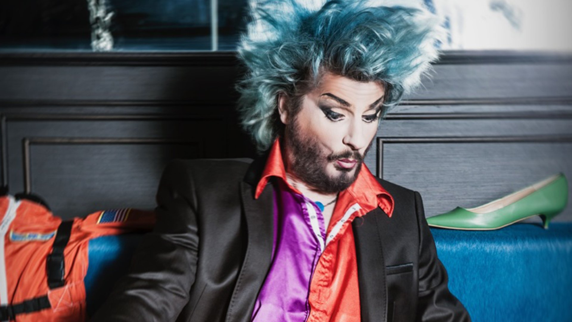 Der Künstler mit blauen Haaren und farbiger Kleidung, nach unten blickend. Rechts im Bild ein grüner Damenschuh