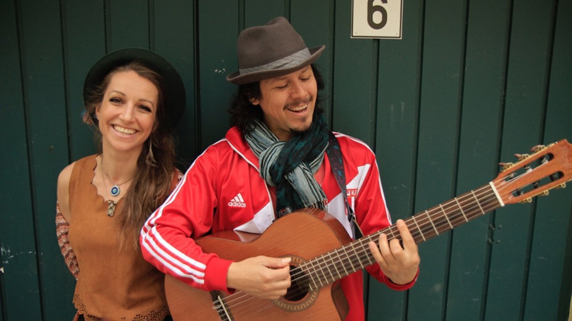 Veranstaltungsfoto eines Live‑Musikauftritts: Zwei Personen stehen vor einer dunkelgrünen Holzwand. Eine Person im Vordergrund spielt eine Akustikgitarre und trägt eine rote Jacke sowie einen Hut. Daneben steht eine weitere Person mit Hut und ärmellosem Oberteil. Die Aufnahme vermittelt eine ungezwungene Atmosphäre eines Singer‑Songwriter‑ oder Akustik‑Konzerts im Freien oder in kleinem Rahmen.
