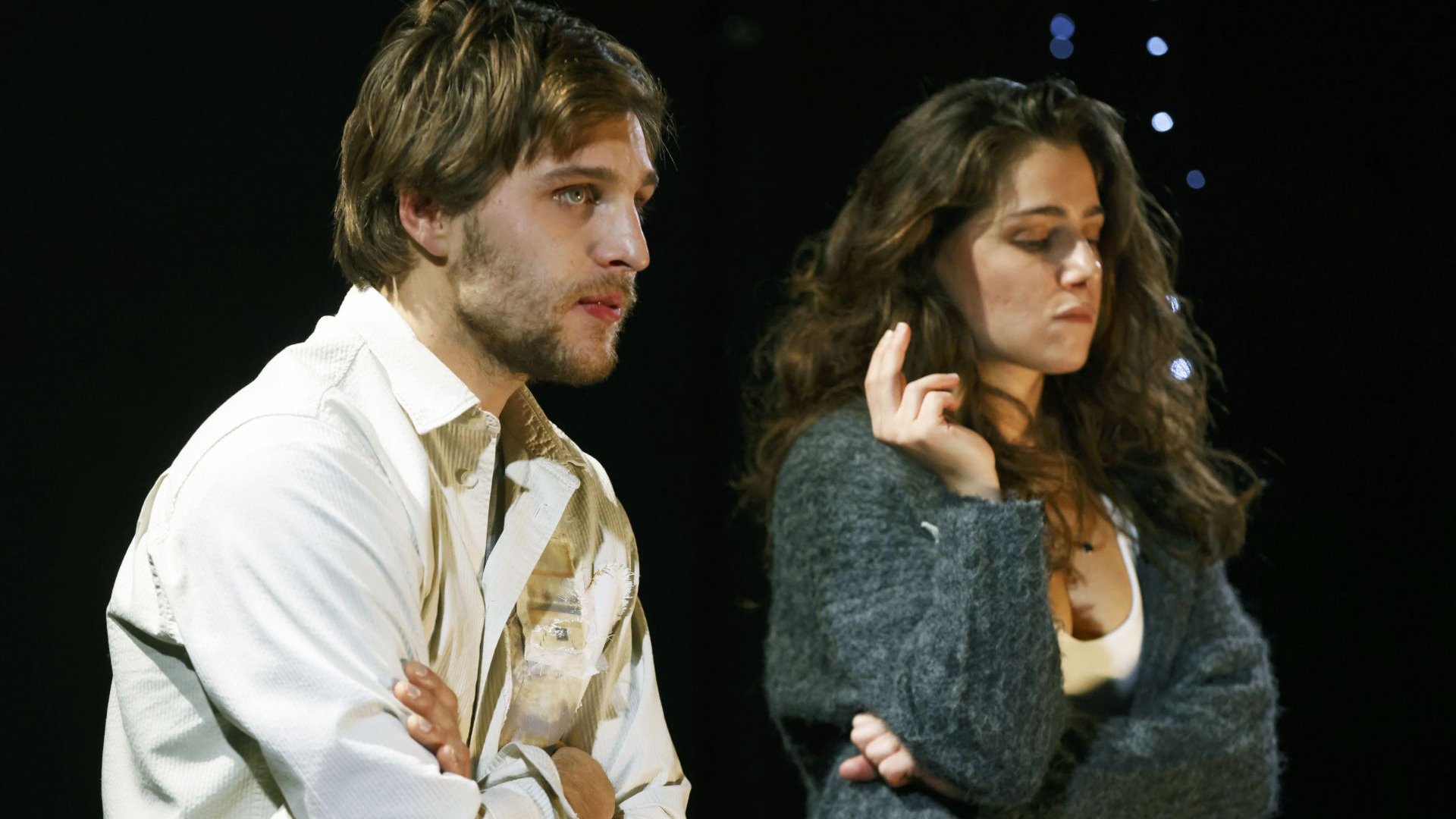 Zwei Personen nebeneinander auf einer Bühne, mit verschränkten Armen, vor dunklem Hintergrund und punktuellem Bühnenlicht