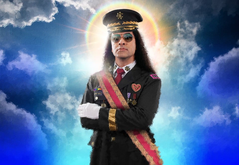 Bülent Ceylan in schwarzer Uniform mit Schirmmütze, Sonnenbrille, Ordensabzeichen und roter Schärpe, vor wolkigem Himmel mit leuchtendem Heiligenschein im Hintergrund.