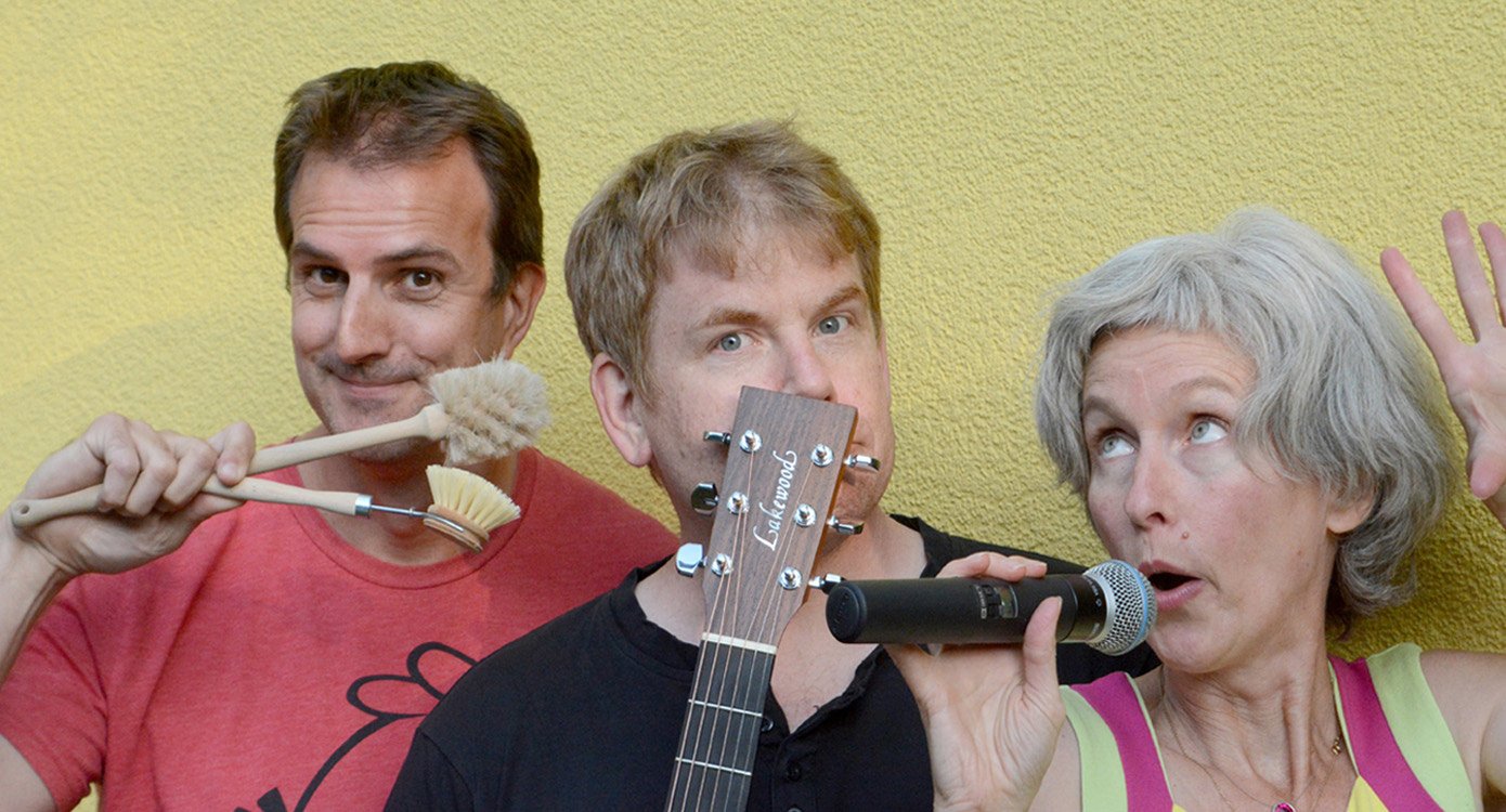 Zwei Männer und eine Frau, sie mit Mikrofon, ein Mann mit Gitarre, ein Mann mit Bürsten vor gelbem Hintergrund