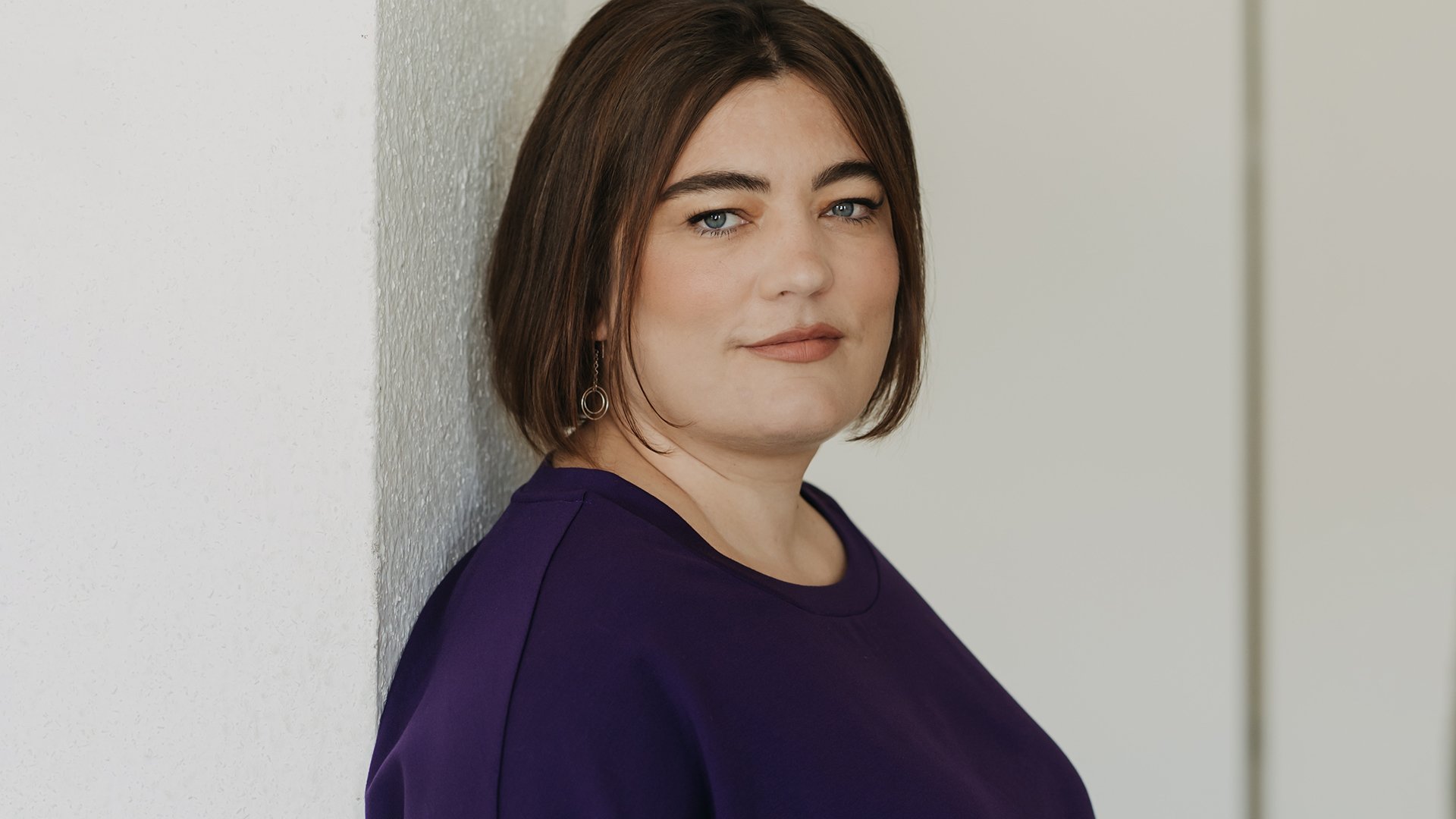 Ein Portrait von Alena Schröder in einem dunkelvioletten Kleid an einer hellen Wand lehnend
