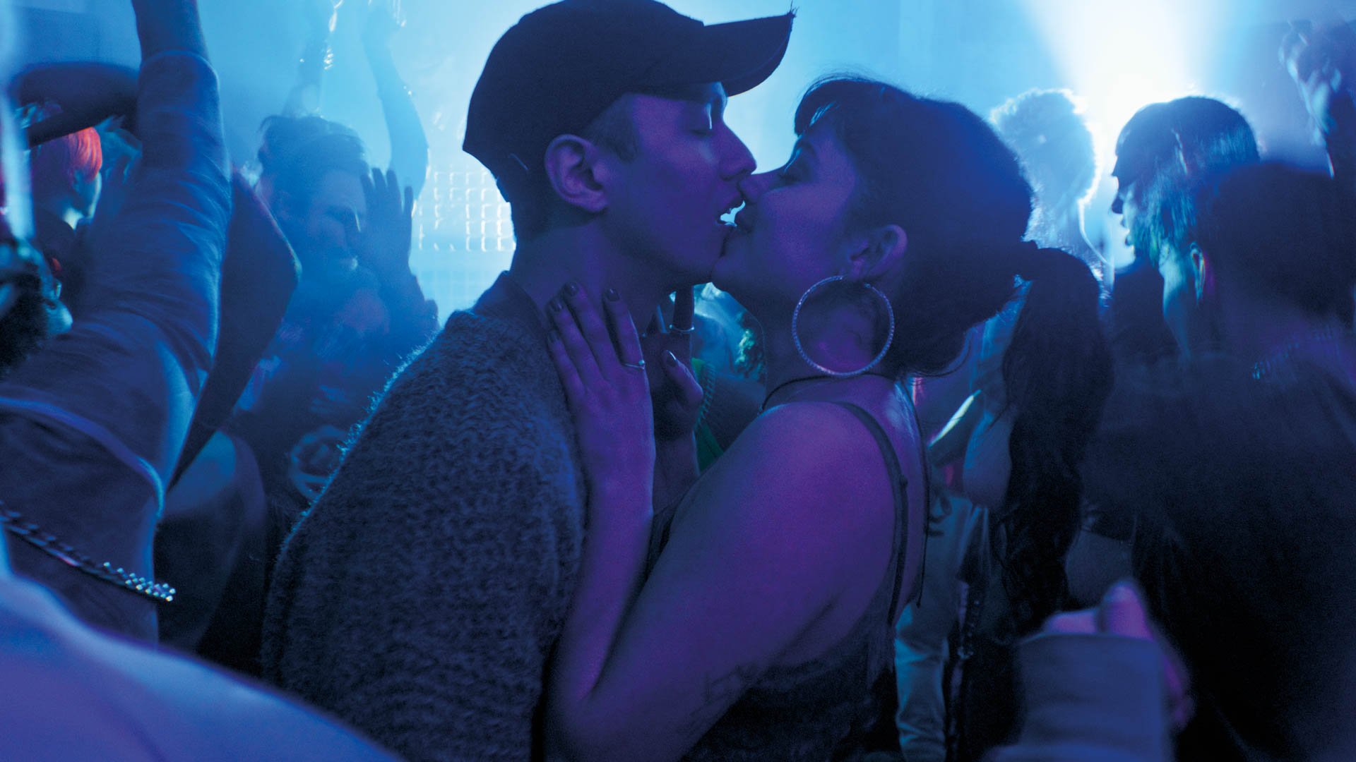 Zwei Personen küssen sich in einem Club, umgeben von tanzenden Menschen und blauem Licht.