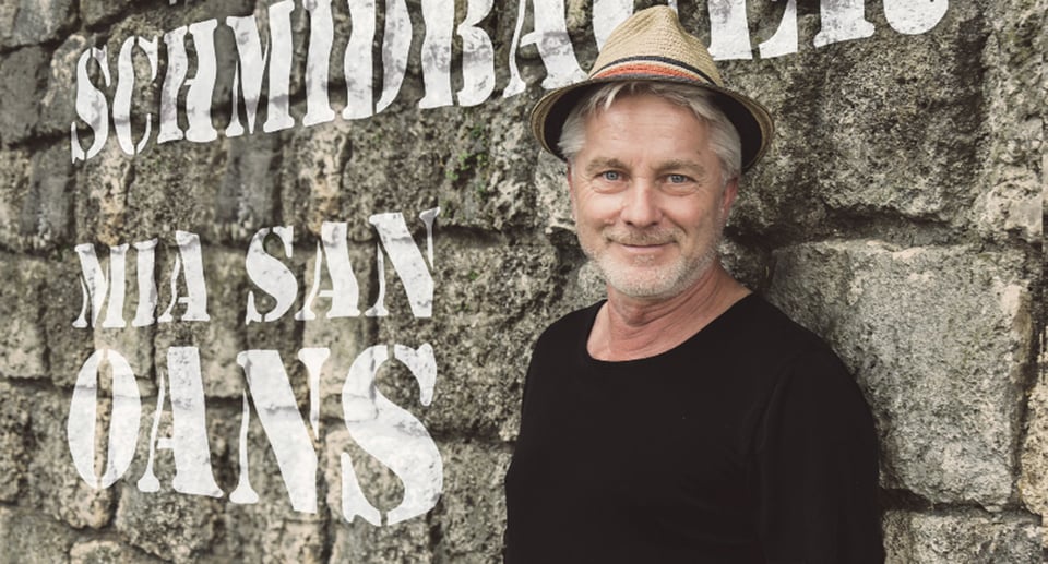 Foto des Künstlers mit schwarzem T-Shirt und Hut auf einer Steinmauer