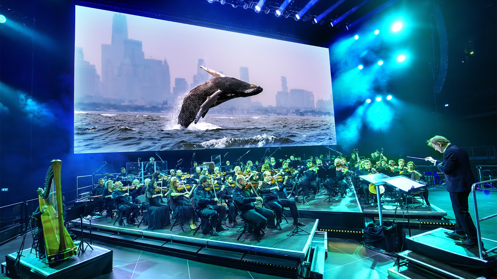 Bühnenfoto eines Symphonieorchesters mit Dirigent auf Podest vor großer Leinwand, die einen aus dem Meer springenden Wal vor einer Stadtkulisse zeigt, umgeben von blauer Bühnenbeleuchtung und Scheinwerfern zur Veranstaltung Unser Blauer Planet III