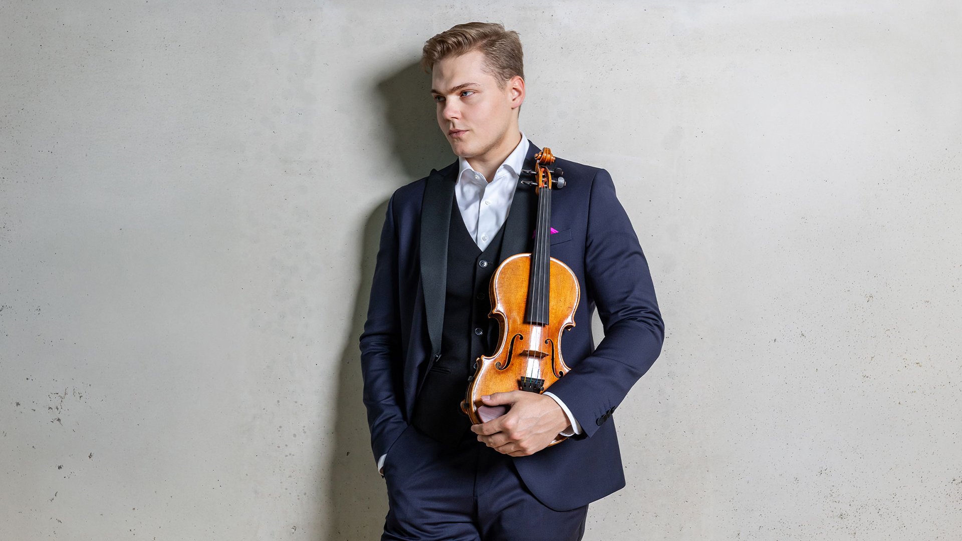 Eventmotiv mit Geiger und Violine: Eine stehende Person in dunklem Anzug hält eine Violine locker vor dem Körper und lehnt vor einer hellen, strukturierten Wand. Der Blick ist seitlich gerichtet, klare Studio‑Beleuchtung hebt Instrument und Kleidung kontrastreich hervor. Reduziertes, elegantes Promofoto für ein klassisches Orchester‑ und Violinkonzert.