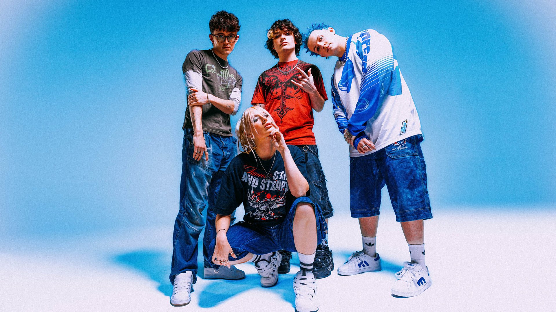 Fünfköpfige Band steht und kniet in einer Studioaufnahme vor blauem Hintergrund, trägt lässige Streetwear in Blau-, Weiß- und Rottönen und posiert gemeinsam für ein Bandfoto.