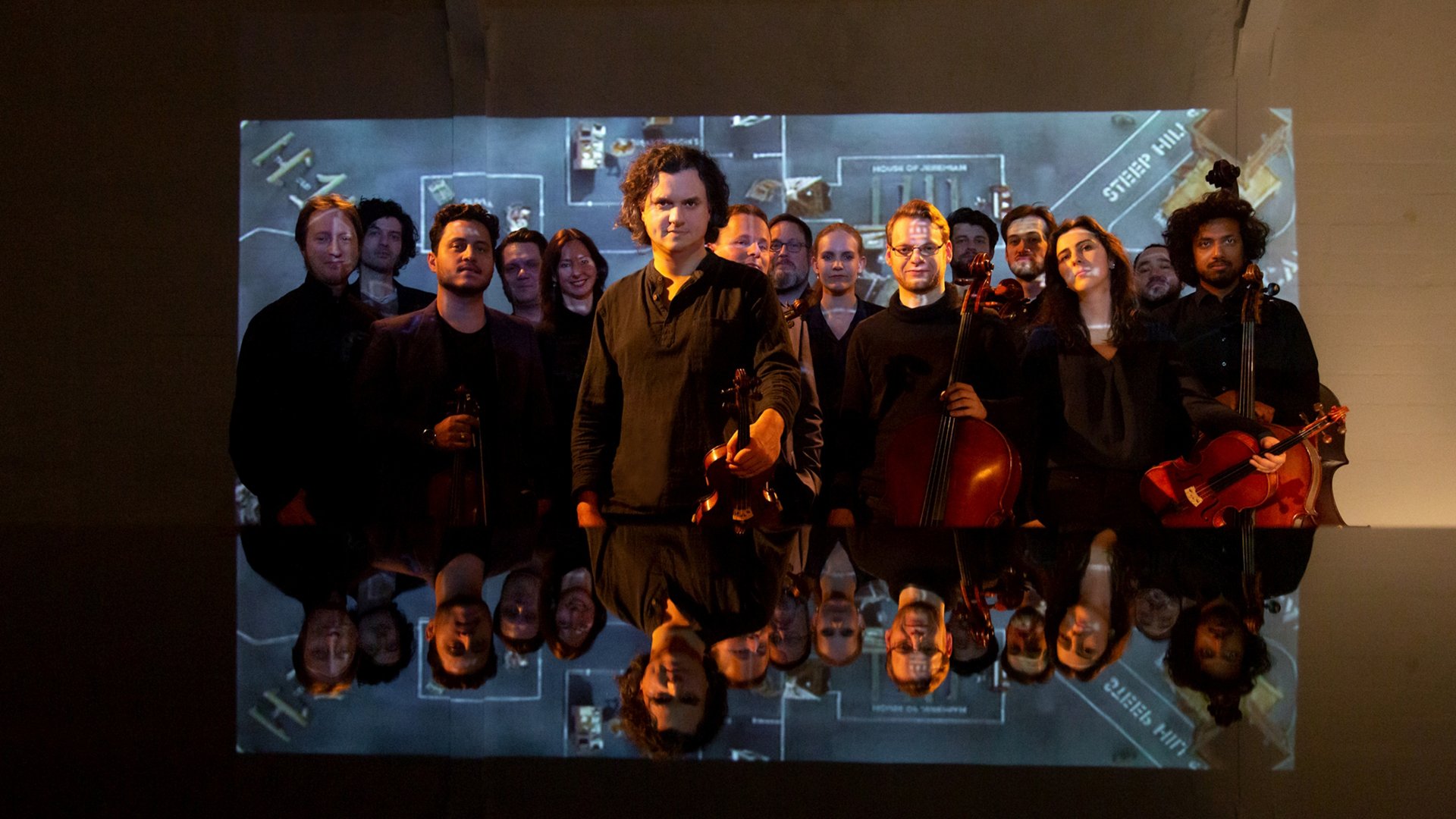 Mitglieder des dogma chamber orchestra mit Streichinstrumenten auf einer Bühne vor einer Videoprojektion.