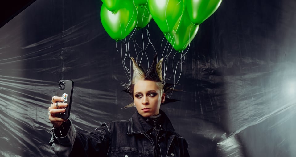 Foto eines Mädchens mit einer modernen, schwarz-blond gefärbten Frisur, in der grüne Luftballons befestigt sind. Sie macht ein Selfie