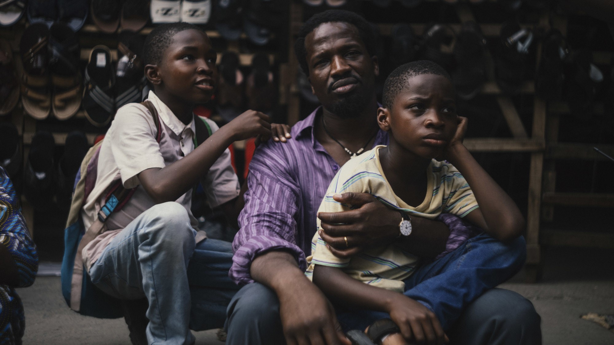Filmszene aus My Father’s Shadow mit einer Person und zwei Kindern, die vor einem Verkaufsstand mit zahlreichen Schuhen auf dem Boden sitzen, umgeben von dicht aufgehängten Schuhpaaren im Hintergrund.